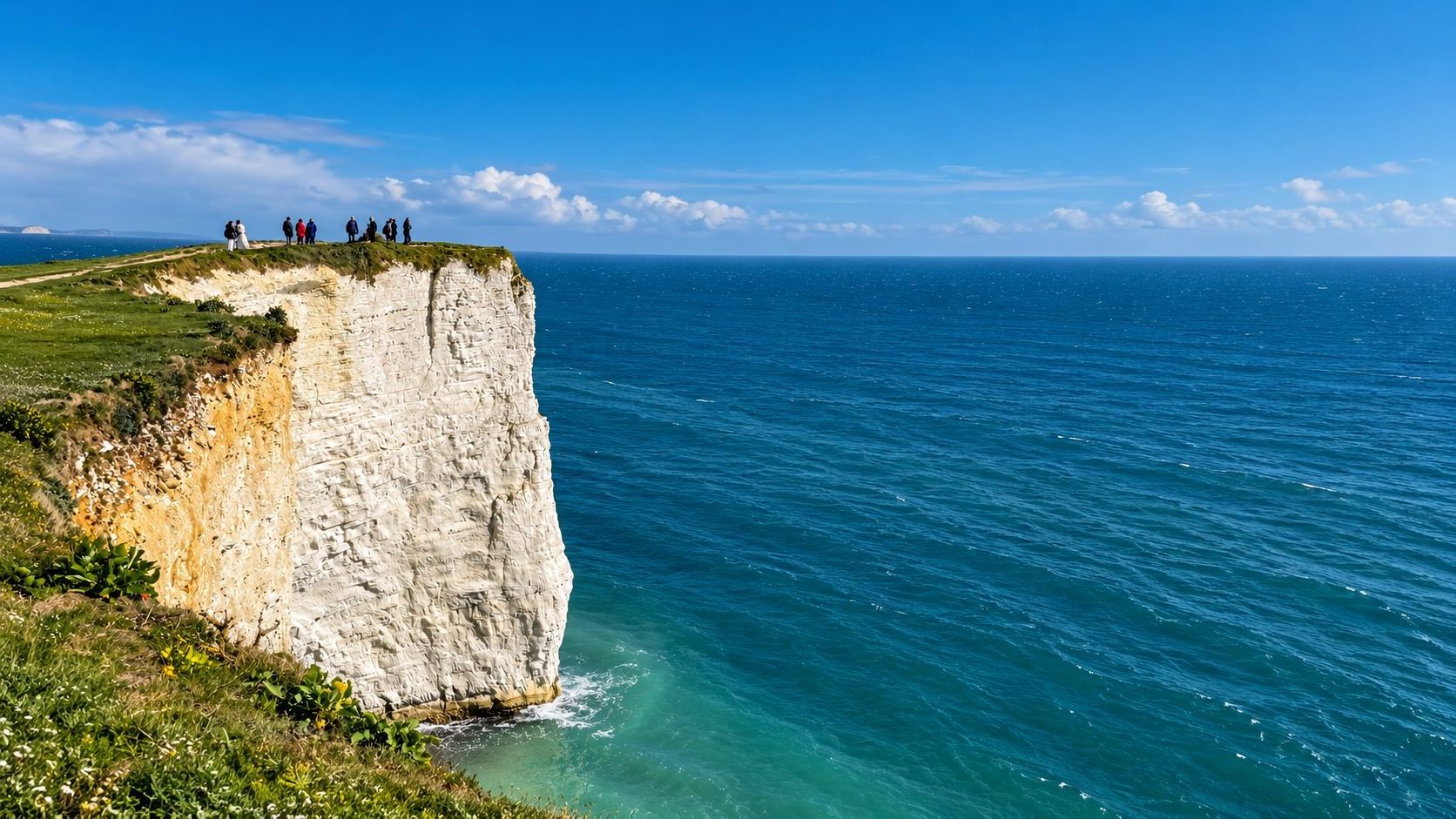 A group of people standing at the top of a sheer white chalk cliff above deep blue sea.